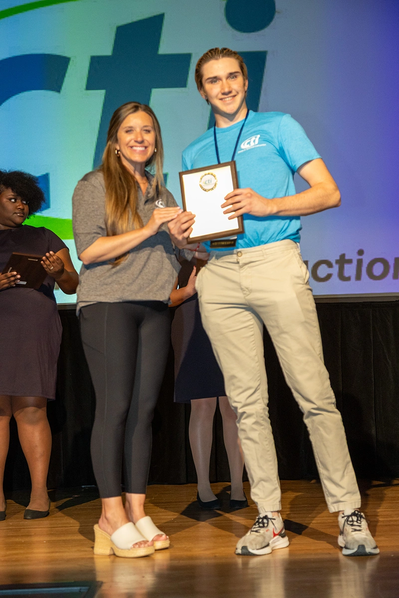 female student on stage in a blue shirt accepting an award from a female adult